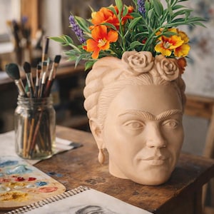 May include: A beige ceramic head sculpture of a woman with a floral crown, filled with colorful artificial flowers. The sculpture is on a wooden table, with art supplies in the background.