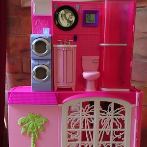 May include: A multi-level dollhouse playset featuring a pink and purple color scheme. The upper level includes a stacked washer and dryer, a vanity with a mirror, a toilet, and a shower. The lower level features a green palm tree and a large window.