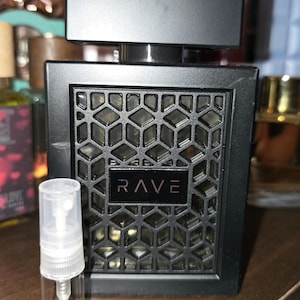 May include: Black rectangular perfume bottle with a geometric pattern and the word "RAVE" in white. A small, clear spray bottle is in front of the perfume bottle. The background is blurred, with other perfume bottles visible.
