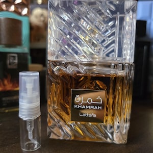 Op de afbeelding: Een rechthoekige parfumfles met een heldere, getextureerde, kristalachtige dop. De fles bevat een goudbruine vloeistof en heeft een gouden label met Arabische en Engelse tekst: "Khamrah" en "Lattafa". Naast de fles staat een kleine, heldere verstuiver.