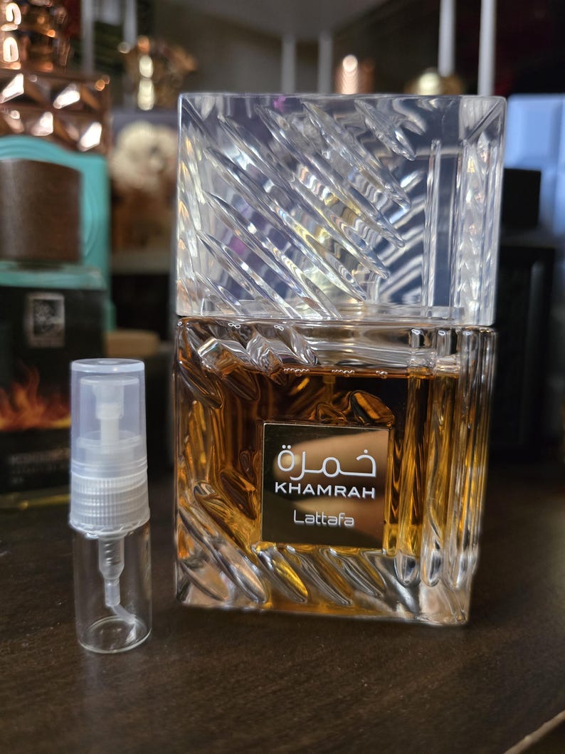 Op de afbeelding: Een rechthoekige parfumfles met een heldere, getextureerde, kristalachtige dop. De fles bevat een goudbruine vloeistof en heeft een gouden label met Arabische en Engelse tekst: "Khamrah" en "Lattafa". Naast de fles staat een kleine, heldere verstuiver.
