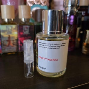 May include: A clear glass perfume bottle with a silver cap and a smaller sample bottle. The label reads "FRUITY NEROLI" and lists fragrance notes. Other perfume bottles are visible in the background.