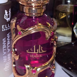May include: A purple perfume bottle with gold filigree and a decorative gold cap. The bottle has the word "HABIK" in gold lettering. The perfume bottle is surrounded by other bottles.
