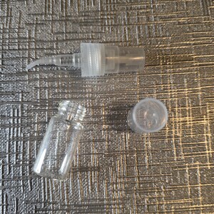Op de afbeelding: Een heldere glazen flacon, een heldere plastic sproeipomp en een heldere plastic dop zijn gerangschikt op een getextureerd zilveren oppervlak. De flacon is cilindrisch met een schroefdraadhals. De pomp heeft een gebogen mondstuk.