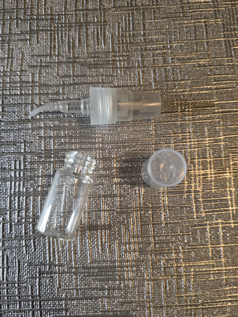 Op de afbeelding: Een heldere glazen flacon, een heldere plastic sproeipomp en een heldere plastic dop zijn gerangschikt op een getextureerd zilveren oppervlak. De flacon is cilindrisch met een schroefdraadhals. De pomp heeft een gebogen mondstuk.