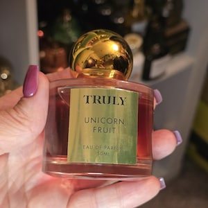 May include: A clear glass perfume bottle with a gold cap is held in a hand. The bottle contains a pink liquid and has a gold label that reads "TRULY UNICORN FRUIT EAU DE PARFUM 50ml."