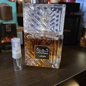 Op de afbeelding: Een heldere, geometrische parfumfles met een getextureerde, schuine bovenkant en een rechthoekige basis. De fles bevat een goudbruine vloeistof en heeft het woord "Khamrah" in Arabisch schrift, samen met "Lattafa" in het Engels. Er staat een kleine, lege spuitfles naast.