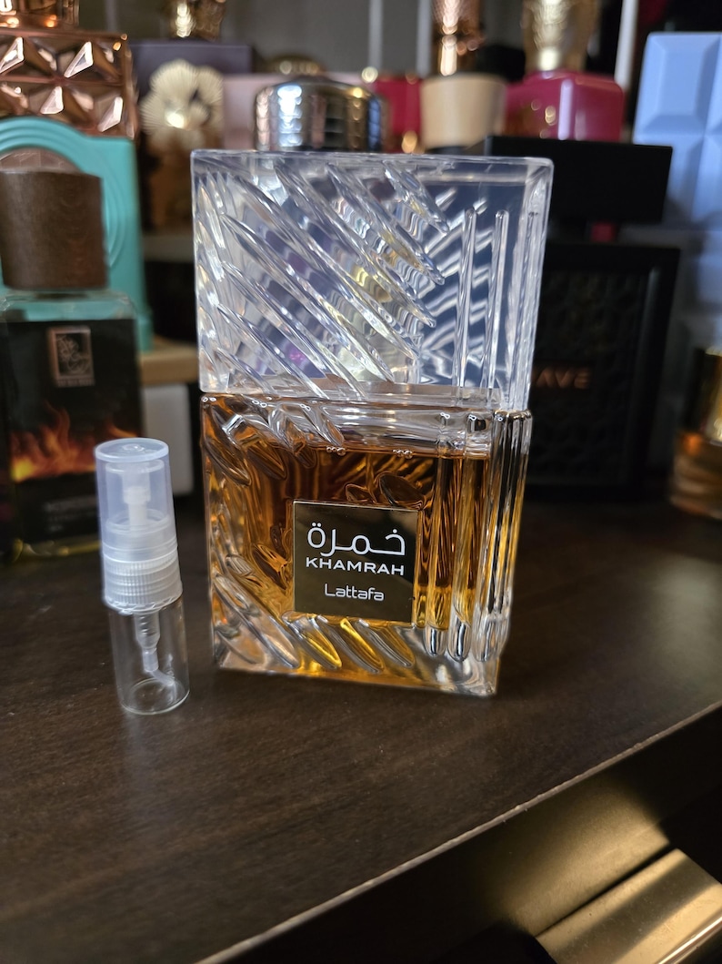 Op de afbeelding: Een heldere, geometrische parfumfles met een getextureerde, schuine bovenkant en een rechthoekige basis. De fles bevat een goudbruine vloeistof en heeft het woord "Khamrah" in Arabisch schrift, samen met "Lattafa" in het Engels. Er staat een kleine, lege spuitfles naast.