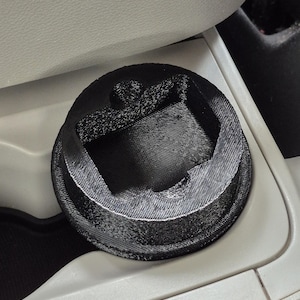 May include: A black, 3D-printed car cup holder insert. The insert has a textured surface and a geometric design. It is designed to fit into a car's existing cup holder.