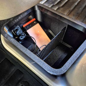 May include: A black car console organizer with a phone, charger, and a carbon fiber accent. The organizer is rectangular with rounded corners and is placed in a car. The phone is peach with an orange top.