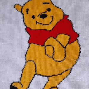 May include: A crocheted blanket featuring a smiling Winnie the Pooh character. The bear is yellow with a red shirt and black outlines, set against a white background. The blanket is handmade with visible stitch detailing.