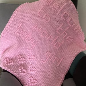 May include: A pink knitted baby blanket with raised lettering and heart motifs. The blanket is draped over a grey chair, highlighting the soft texture and detailed craftsmanship of the handmade item.