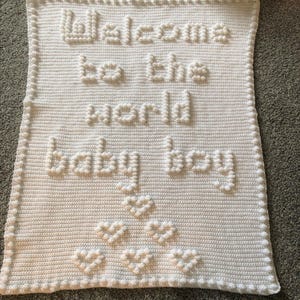 May include: A white crocheted baby blanket with the text "Welcome to the world baby boy" in raised letters. The blanket also features raised heart shapes. The edges are finished with a decorative border.