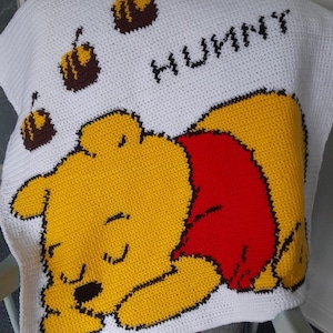 Könnte beinhalten: Eine weiße Häkeldecke mit einem gelben Zeichentrickbären mit rotem Hemd und dem Wort "HUNNY" in Schwarz. Die Decke hat auch braune und gelbe Honigtopf-Designs.