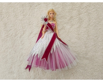 May include: A doll with blonde hair wearing a floor-length gown featuring a white and silver glitter bodice, a layered tulle skirt in shades of pink and white, and a deep burgundy velvet sash draped across the front with a silver brooch.