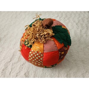 May include: A stuffed fabric pumpkin decoration featuring a patchwork design of red, orange, and brown fabrics with various patterns including gingham and checks. It has dark green fabric leaves and a brown stem, topped with a cluster of tan raffia.