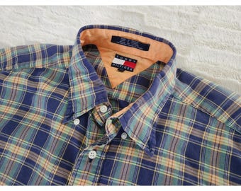May include: A button-down plaid shirt in shades of blue, green, and tan. The shirt features a button-down collar and a small Tommy Hilfiger logo. The collar lining is a peach color.
