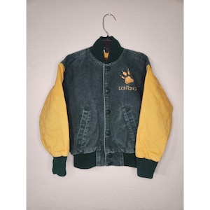 Puede incluir: Chaqueta vaquera verde oscuro con mangas amarillas. La chaqueta presenta una huella de pata y el texto "The Lion King" en dorado. Tiene cierre de botones, cuello verde y puños verdes. La chaqueta está colgada de una percha metálica.