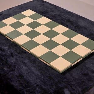 May include: A rectangular chessboard with alternating green and beige squares. The board is set on a dark blue surface, creating a contrast with the lighter colours of the board. The squares are evenly sized and arranged in a classic checkerboard pattern.