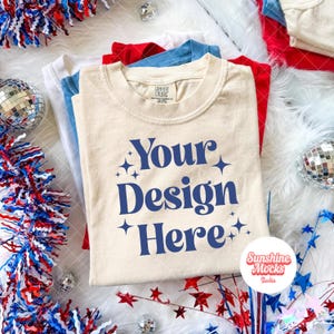 Peut inclure: Une pile de t-shirts rouges, blancs et bleus, avec un t-shirt crème sur le dessus. Le t-shirt crème porte le texte "Your Design Here" en bleu. Des boules disco et des ornements en forme d'étoiles décorent l'ensemble.