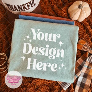 May include: A sage green t-shirt featuring the white text Your Design Here surrounded by four-pointed star icons. The shirt is folded and stacked with other shirts in blue and orange, accompanied by a small white pumpkin and a plaid ribbon.