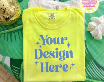 Op de afbeelding: Twee T-shirts, één witte en één neongele, worden gepresenteerd op een oppervlak met een patroon. Het gele shirt heeft de blauwe tekst "Your Design Here". Een discobal en een scrunchie zijn ook zichtbaar.