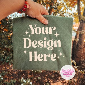 May include: A sage green t-shirt featuring the cream-colored text "Your Design Here" in a bold, serif font. The text is surrounded by small, four-pointed star icons. The fabric has a soft, matte texture and is held up against a blurred outdoor background.