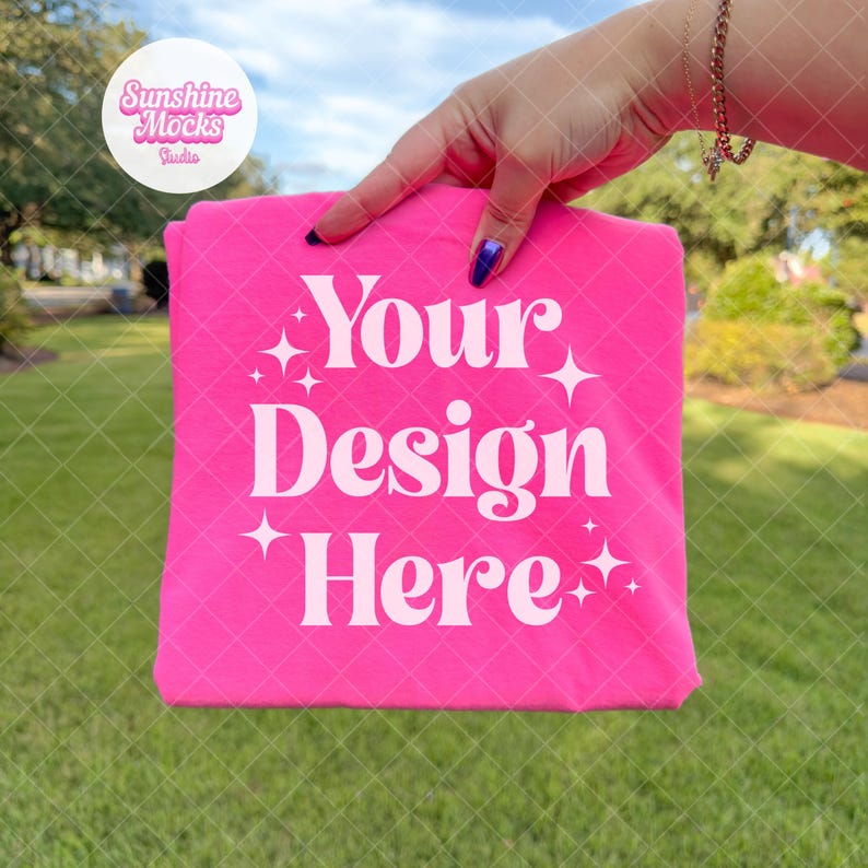 K&ouml;nnte beinhalten: Ein leuchtend pinkfarbenes T-Shirt mit dem wei&szlig;en Schriftzug "Your Design Here" in stilisiertem Text. Das Shirt wird vor einem gr&uuml;nen Gras Hintergrund gehalten. Sternf&ouml;rmige Grafiken umgeben den Text.