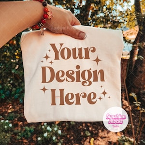 Op de afbeelding: Een crèmekleurig T-shirt met de bruine tekst Your Design Here, omringd door kleine vierpuntige sterpictogrammen. Het shirt wordt vastgehouden tegen een wazige buitenachtergrond, waardoor de centrale grafische print duidelijk zichtbaar is.