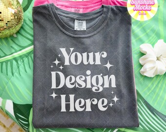May include: A dark gray t-shirt with the text "Your Design Here" in white, layered over a pink t-shirt. The shirts are on a green and white patterned surface, with a disco ball and a white scrunchie in the background.