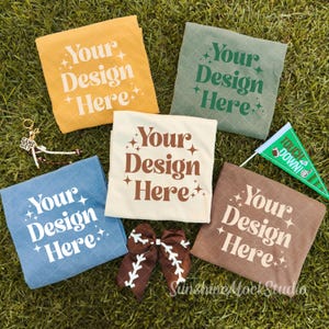Puede incluir: Cinco camisetas dobladas en amarillo mostaza, verde salvia, crema, azul y marrón, cada una con el texto Your Design Here y estrellas. Incluye un lazo para el pelo marrón con costuras blancas, un llavero y un banderín verde con Touchdown.