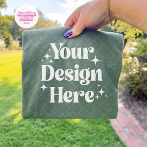 Op de afbeelding: Een opgevouwen, olijfgroene stof met de witte tekst "Your Design Here". De stof wordt vastgehouden door een hand met een gouden armband en paarse nagellak. De achtergrond is wazig, wat duidt op een buitenomgeving.