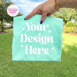 May include: A folded, light turquoise t-shirt with the text "Your Design Here" in white. The shirt is held up by a hand with a gold bracelet and purple nail polish. The background is a green lawn and trees.