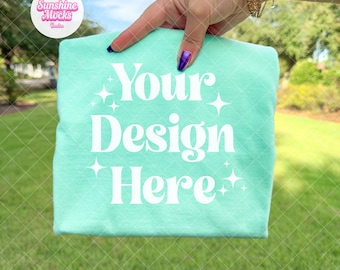May include: A folded, light turquoise t-shirt with the text "Your Design Here" in white. The shirt is held up by a hand with a gold bracelet and purple nail polish. The background is a green lawn and trees.