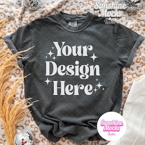 May include: Dark gray t-shirt with the text "Your Design Here" in white, surrounded by starbursts. The shirt is displayed on a patterned blanket with decorative elements, including disco balls and pampas grass. A white boot is visible.
