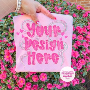 May include: A light pink sweatshirt with the text "Your Design Here" in a bubble font. The design includes pink hearts and swirls. The sweatshirt is held up by a hand with a bracelet and pink nail polish. The background is a bush with pink flowers.