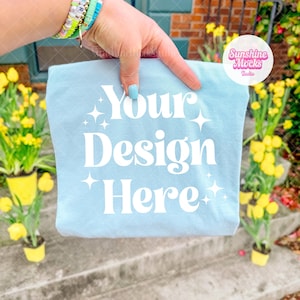 May include: A light blue t-shirt with the text "Your Design Here" in white. The shirt is held up by a hand with bracelets. The background includes yellow flowers in pots and a brick wall.