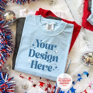May include: A stack of folded t-shirts in various colors, including light blue, red, white, and charcoal gray. The light blue shirt features the text "Your Design Here" in a dark blue font. Decorative elements include disco balls and patriotic-themed tinsel.