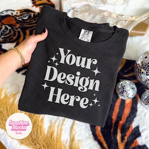 Image of a girl holding up a folded Black Comfort Colors lightweight sweatshirt with rhinestone cowgirl boots, mirrored disco balls, and pampas grass. There is of a tiger blanket in the background. The style is boho, western, and preppy for an aesthetic trendy look.