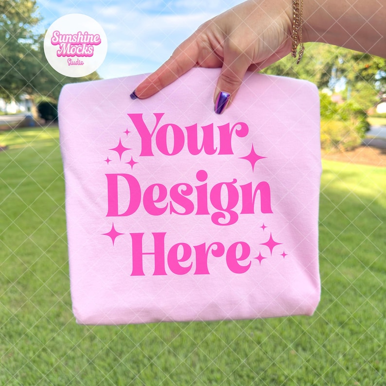 May include: A light pink sweatshirt with the text "Your Design Here" in bright pink. The sweatshirt is held up by a hand with purple nail polish, against a green grass background.