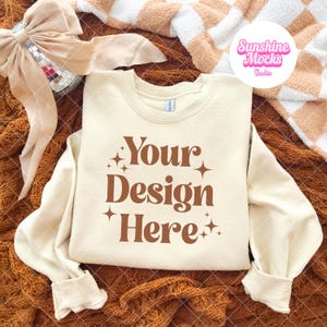 Könnte beinhalten: Cremefarbenes Sweatshirt mit dem Text "Your Design Here" in Braun. Das Sweatshirt wird auf einer braunen Strickoberfläche präsentiert, mit einer beigen Schleife und einer Discokugel im Hintergrund. Eine weiß-beige karierte Decke ist ebenfalls zu sehen.