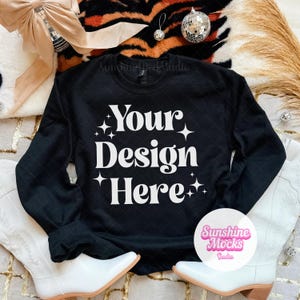 May include: Black crewneck sweatshirt with the text "Your Design Here" in white. The sweatshirt is displayed on a patterned surface with white cowboy boots and decorative elements.