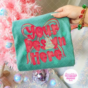 May include: Teal t-shirt with the text "Your Design Here" in red bubble letters, accented with a rainbow design and snowflakes. A small Christmas tree graphic is in the center. The shirt is held up in front of a pink Christmas tree.