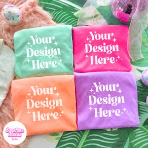 May include: Four folded t-shirts in shades of mint green, hot pink, orange, and purple. Each shirt displays the text "Your Design Here" in white, with star accents. The shirts are arranged on a textured surface with decorative elements.
