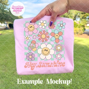 May include: A light pink t-shirt featuring a colorful floral design with smiley faces and the text "Hey Sunshine." The design includes daisies, a peace sign, and a checkered background. The words "Example Mockup!" are at the bottom.