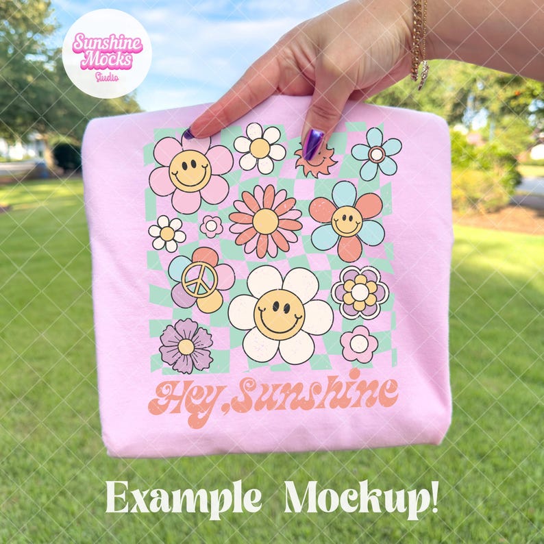 May include: A light pink t-shirt featuring a colorful floral design with smiley faces and the text "Hey Sunshine." The design includes daisies, a peace sign, and a checkered background. The words "Example Mockup!" are at the bottom.