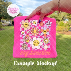 K&ouml;nnte beinhalten: Ein leuchtend pinkfarbenes T-Shirt mit einem Retro-Blumenmuster, einschlie&szlig;lich Smiley-Gesichtern und Friedenssymbolen, und dem Text "Hey Sunshine". Das Design ist vor einem karierten Hintergrund platziert. Das T-Shirt wird vor einem gr&uuml;nen, grasbewachsenen Hintergrund pr&auml;sentiert.