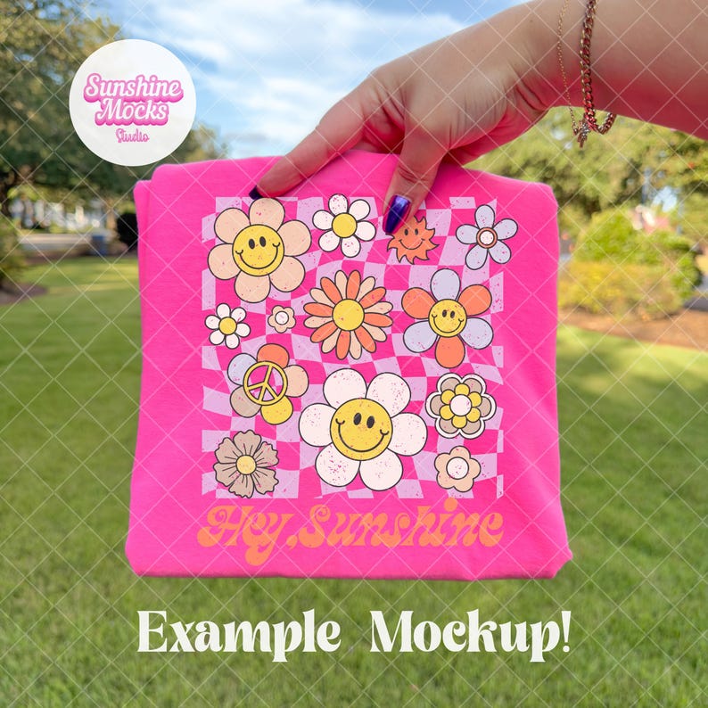 K&ouml;nnte beinhalten: Ein leuchtend pinkfarbenes T-Shirt mit einem Retro-Blumenmuster, einschlie&szlig;lich Smiley-Gesichtern und Friedenssymbolen, und dem Text "Hey Sunshine". Das Design ist vor einem karierten Hintergrund platziert. Das T-Shirt wird vor einem gr&uuml;nen, grasbewachsenen Hintergrund pr&auml;sentiert.