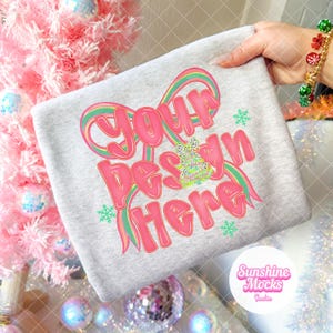 Könnte beinhalten: Ein gefaltetes graues T-Shirt mit dem Text "Your Design Here" in Pink, mit einem Regenbogen-Schleifen-Design. Das Shirt hat auch eine kleine Weihnachtsbaum-Grafik und Schneeflocken. Das Sunshine Mocks Studio-Logo befindet sich unten rechts.