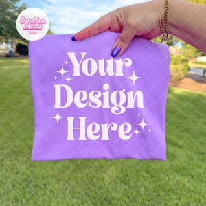 May include: A lavender t-shirt with the text "Your Design Here" in white, with star accents. The shirt is held up against a background of green grass and a blurred outdoor scene. The image includes a logo in the upper left corner.